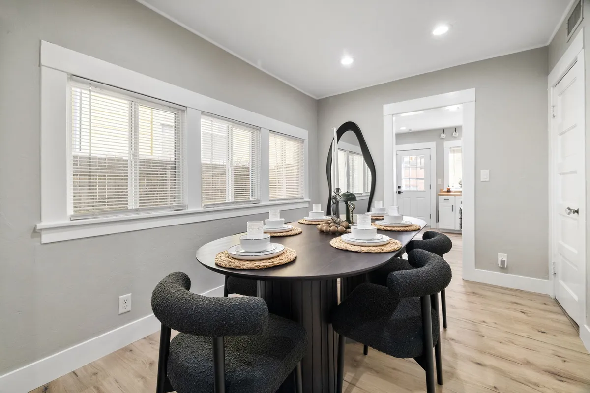 A round dining table surrounded by stylish chairs, accented by a decorative mirror and a sunlit window with blinds. The nook is bright and inviting, ideal for shared meals or relaxed brunches.