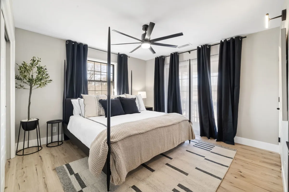 This image features a serene bedroom with a bed adorned in decorative pillows, large windows draped with curtains allowing natural light to pour in, a ceiling fan, and an indoor plant. The overall ambiance is calm and inviting, ideal for relaxing, reading, or sleeping.