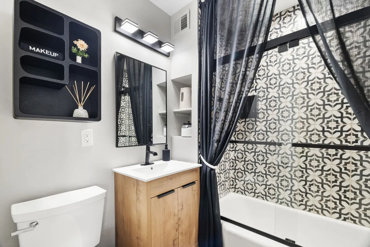 A modern bathroom featuring a vanity with a stylish sink, decorative mirror, a shower with a patterned curtain, and a makeup storage shelf. The space feels bright, contemporary, and spa-like, perfect for unwinding after a day out.