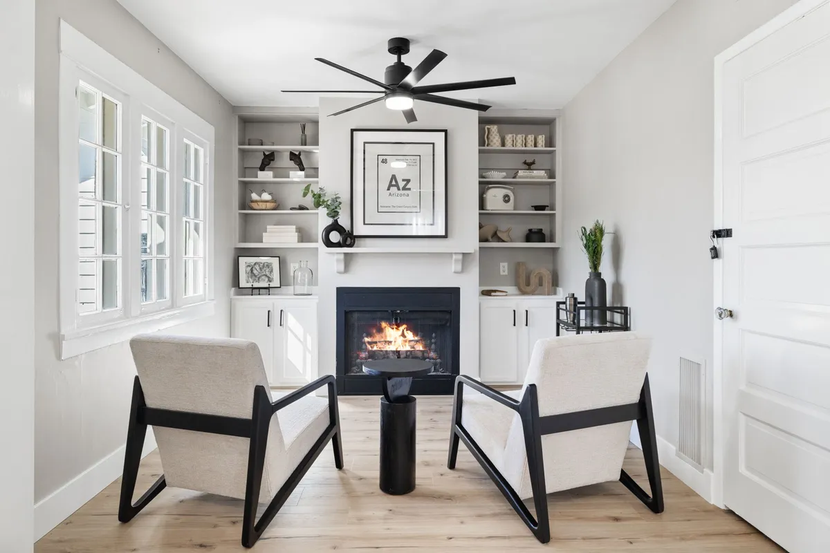A warmly lit sitting room featuring a prominent fireplace as the centerpiece, surrounded by an inviting seating arrangement. Shelves adorned with period-appropriate décor flank the fireplace, and natural light spills into the space, highlighting the restored woodwork and cozy ambiance.