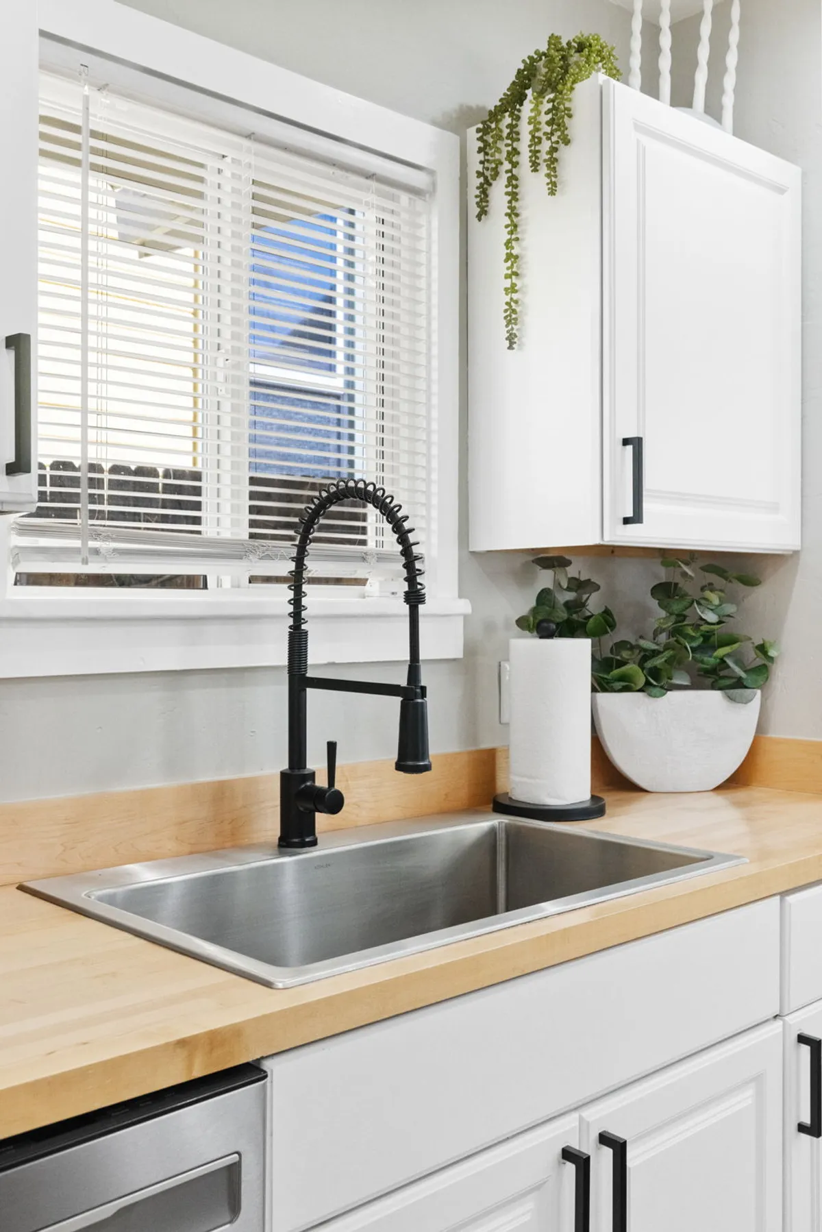 This image features a stylish kitchen area with a black faucet, stainless steel sink, and a wooden countertop adorned with potted plants. The setup feels fresh and modern, suggesting an inviting space for preparing coffee or meals. The presence of natural light and greenery enhances the contemporary urban vibe.